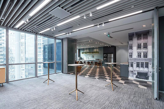 Modern office lounge area with standing tables, kitchen, and large windows overlooking skyscrapers.