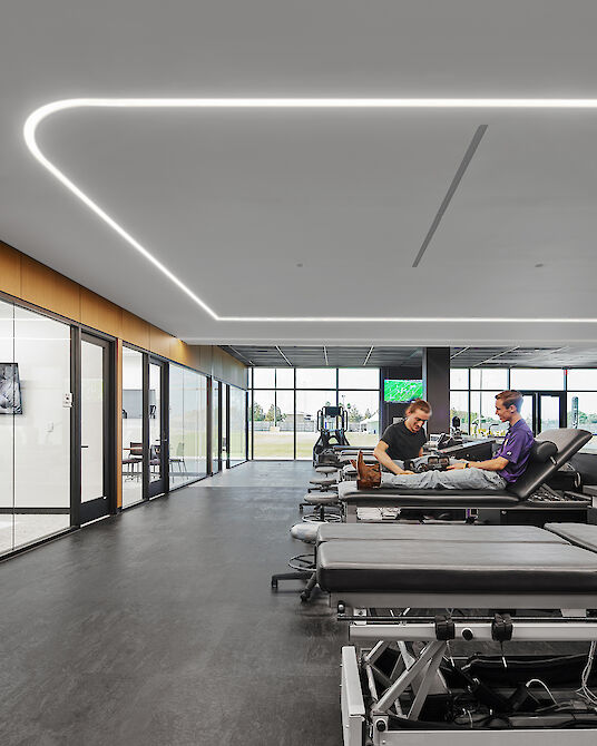 Light-filled rehabilitation room with treatment beds, LED strip lighting and a view of the outdoor playing field