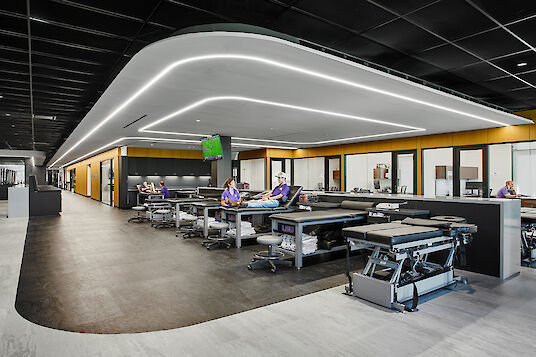 Open rehabilitation area with several treatment beds, LED strip lighting and glass office units in the background
