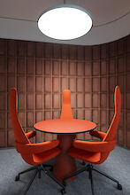 Round conference table with three tall red chairs under a round pendant lamp in front of a brown wall