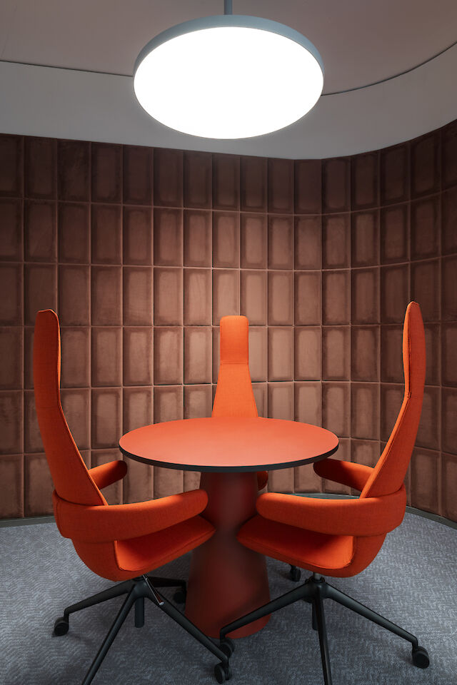 Round conference table with three tall red chairs under a round pendant lamp in front of a brown wall