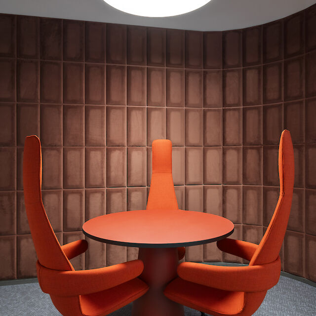 Round conference table with three tall red chairs under a round pendant lamp in front of a brown wall