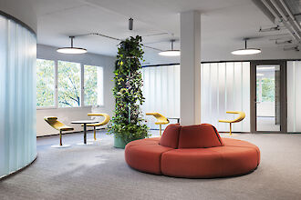 Bright lounge office with red round sofa, yellow chairs, green plant wall and round pendant lights