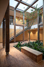 Cour intérieure du Håndværkskollegiet avec sol en briques, constructions en bois et espace vert avec arbre ; la lumière du jour pénètre par un toit vitré, des projecteurs Midpoint éclairent les murs et la pièce.