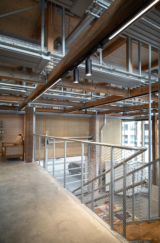Vista interior de una zona de escaleras en el Håndværkskollegiet con vigas de madera a la vista, estructuras metálicas y dos focos Midpoint negros de Lightnet en el techo.