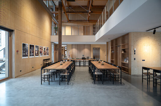 Grande salle commune au Håndværkskollegiet avec de longues tables en bois et des chaises noires ; murs et plafond en bois, sol en béton et spots Midpoint de Lightnet pour l'éclairage