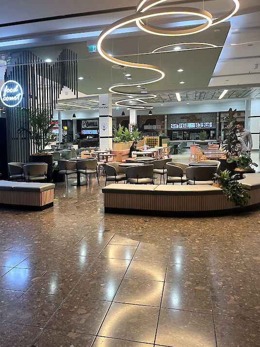 Bright food court with round seating islands, plants, and curved Liquid Line LED ceiling lights.