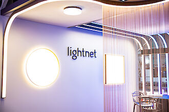 Beleuchteter Lightnet-Messestand mit runder Wand­leuchte, Linienlicht und Sitzbereich hinter Kettenvorhang.