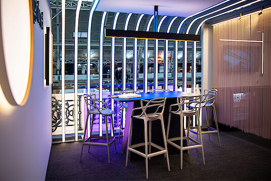 Modern beleuchteter Messestand mit hohen Stühlen, Tisch und vertikalen Lichtstrukturen im Hintergrund.