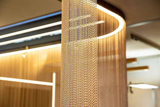 Goldfarbener Kettenvorhang mit integrierter Lichtlinie, erzeugt warmes, strukturiertes Lichtspiel im Raum