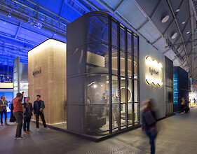 Messestand mit Lichtinstallation, Metallstruktur und beleuchtetem Kubus, Besucher bewegen sich im Raum