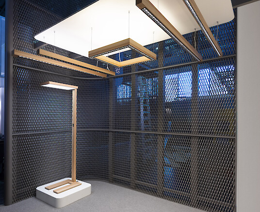 Messestand mit Metallgitterwänden, linearen Holzleuchten und freistehender Stehleuchte auf Podest