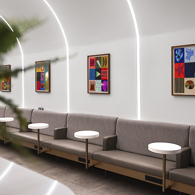 A lounge with benches, small tables, artwork, and curved recessed lights along the wall