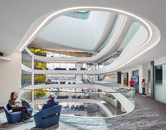 An open atrium with curved levels, lines of light, green walls, and people sitting and walking