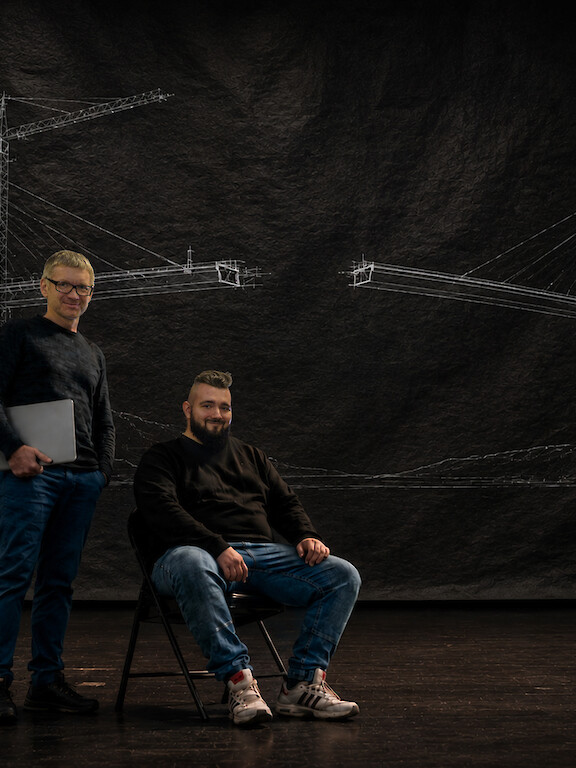 Two men against a dark backdrop with a technical drawing of a bridge structure.