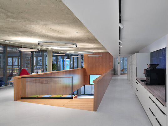 Espacio interior diáfano con galería, barandilla de madera, cocina americana y lámparas colgantes en forma de anillo bajo el techo de hormigón