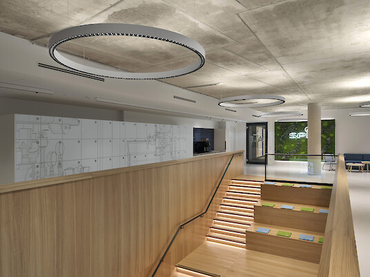 Escalera con revestimiento de madera, peldaños con asiento, barandilla de cristal y lámparas colgantes circulares bajo el techo de hormigón