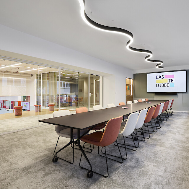 Sala de reuniones con una mesa alargada, sillas, una pared de cristal y una lámpara colgante curvada sobre la mesa