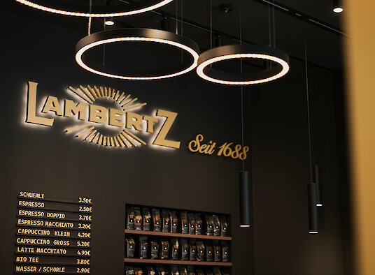 Lambertz-Schriftzug mit Ringpendelleuchten und schlanken Pendelstrahlern vor dunkler Wand