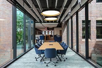 A meeting area with a large wooden table, blue chairs, and round pendant lights set between glass facades