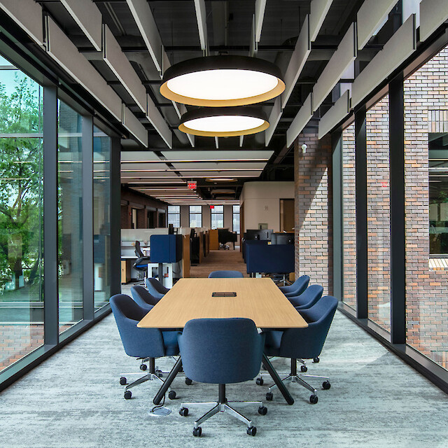 A meeting area with a large wooden table, blue chairs, and round pendant lights set between glass facades