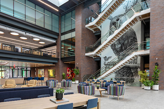 A bright office atrium with an open staircase, seating areas, wall art, and round light fixtures on the upper floor