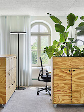 A workspace with a floor lamp, large plants, wooden furniture and a arched window in a bright office