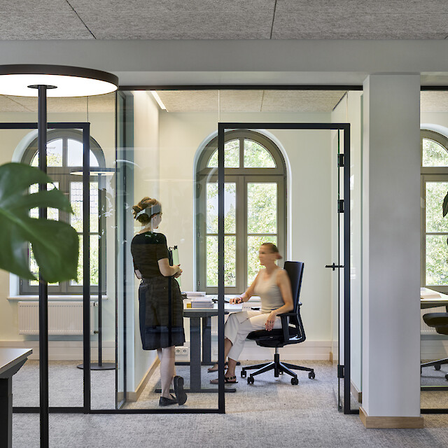 Despachos individuales acristalados con empleados, lámparas de pie, plantas y grandes ventanas con arcos de medio punto que dejan pasar la luz natural