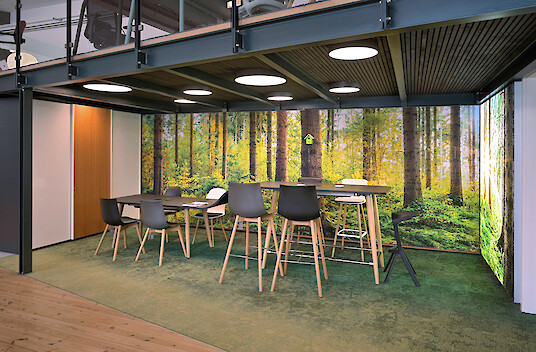 Meeting area with a high table, forest-themed decor, round ceiling lights, and a gallery level above