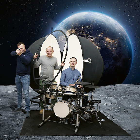 Three men with a drum kit and large luminaire against a staged lunar landscape.