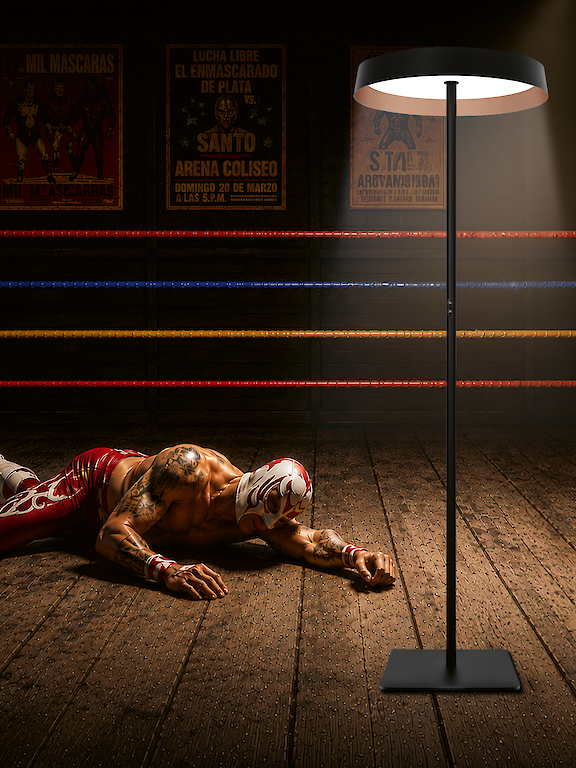 Masked wrestler lying in a boxing ring, illuminated by a floor lamp.