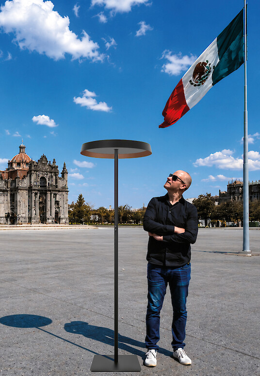 Man standing beside a floor lamp in front of a cathedral and Mexican flag.