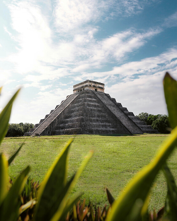 Historic Maya pyramid rising above a green lawn beneath a cloudy sky.