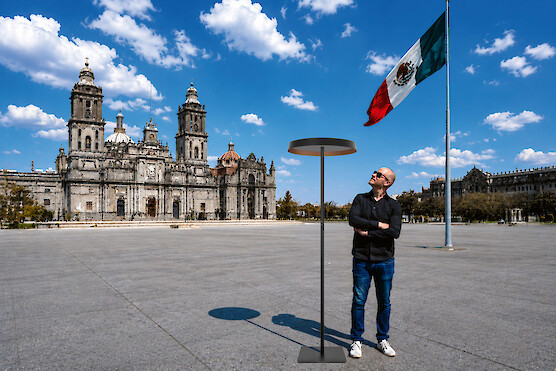Man standing beside a floor lamp in front of a cathedral and Mexican flag.