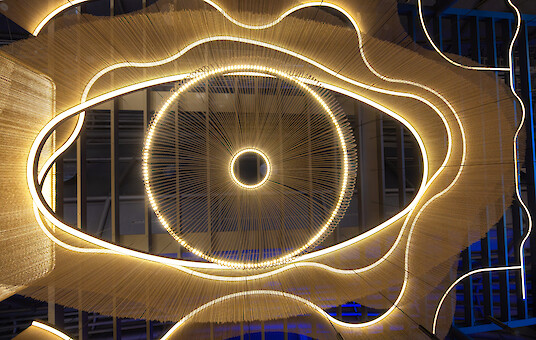 Glowing, curved lines of light and golden chains form an organic ceiling installation with a circular center.