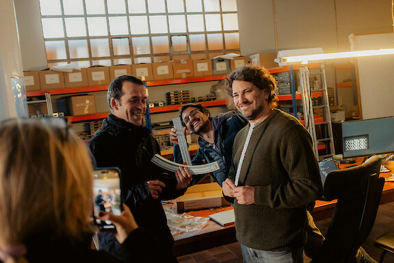 Three men laughing while examining curved lighting elements in a workshop.