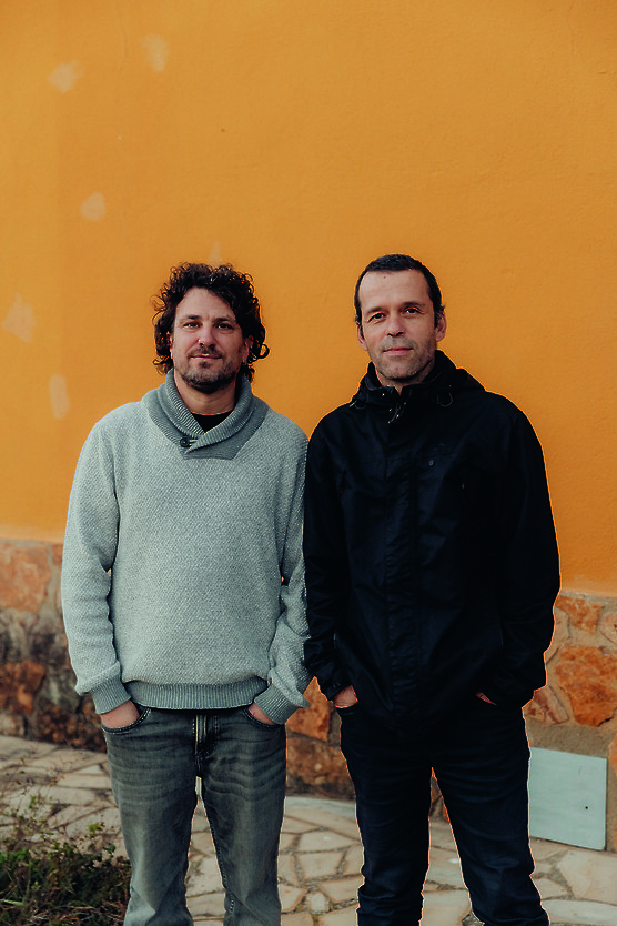 Two men standing side by side in front of an orange wall.
