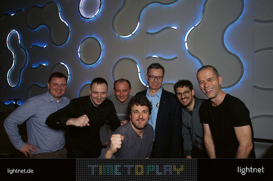 Six men posing together in front of a wall with glowing blue lines.