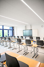 Bright classroom with rows of desks, black chairs and long linear pendant luminaires.