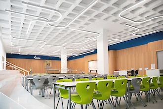 Spacious canteen with green chairs and curved linear pendant luminaires across the ceiling.