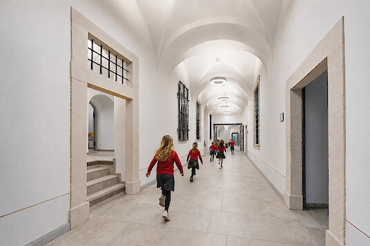 Children walk through a bright school hallway with high vaulted ceilings, a stone floor, and ring-shaped ceiling luminaires.