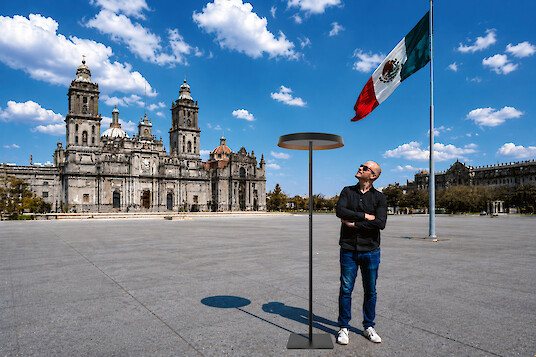 Man standing beside a floor lamp in front of a cathedral and Mexican flag.