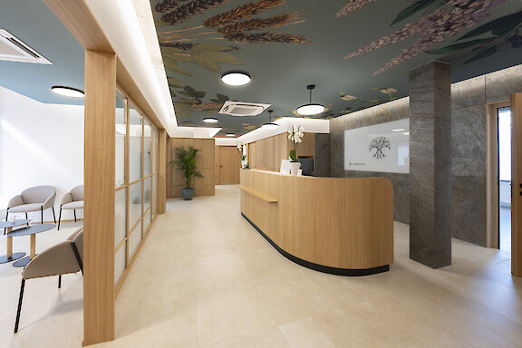 Reception at the Baldi medical practice in Basel with a wooden counter, a ceiling featuring a floral design, and round ceiling and pendant luminaires by Lightnet.
