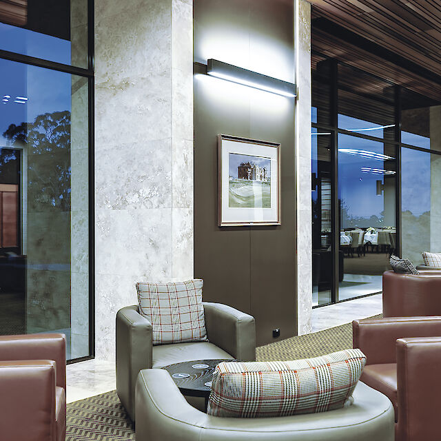 Salón con sillones de cuero, iluminación de pared, columnas de mármol y grandes ventanales con vistas al exterior.