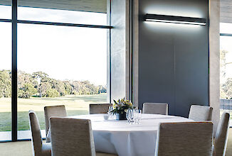 Restaurante con mesa redonda, sillas grises, luz de pared directa/indirecta y ventanas panorámicas con vistas a la naturaleza.