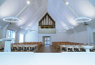 Helle Kirche mit hohen, weißen Decken und ringförmigen Pendelleuchten, symmetrisch angeordnet über Sitzreihen.
