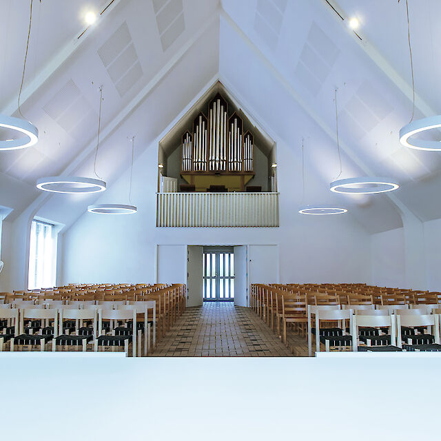 Helle Kirche mit hohen, weißen Decken und ringförmigen Pendelleuchten, symmetrisch angeordnet über Sitzreihen.