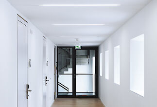 Heller Flur mit linearer LED-Deckenbeleuchtung, weißen Wänden, Türen und Glaszugang zu einer Treppe.