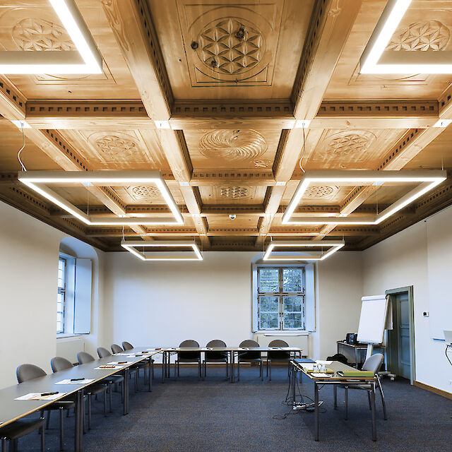 Conference room with ornate wooden ceiling and modern, rectangular LED pendant luminaires above tables arranged in a U-shape.