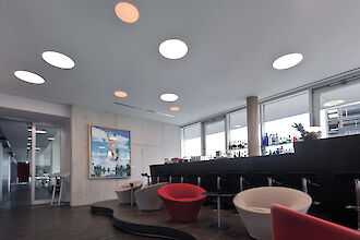 Salón con luminarias redondas empotradas en el techo, elegantes sillones y una barra oscura con frente de cristal.